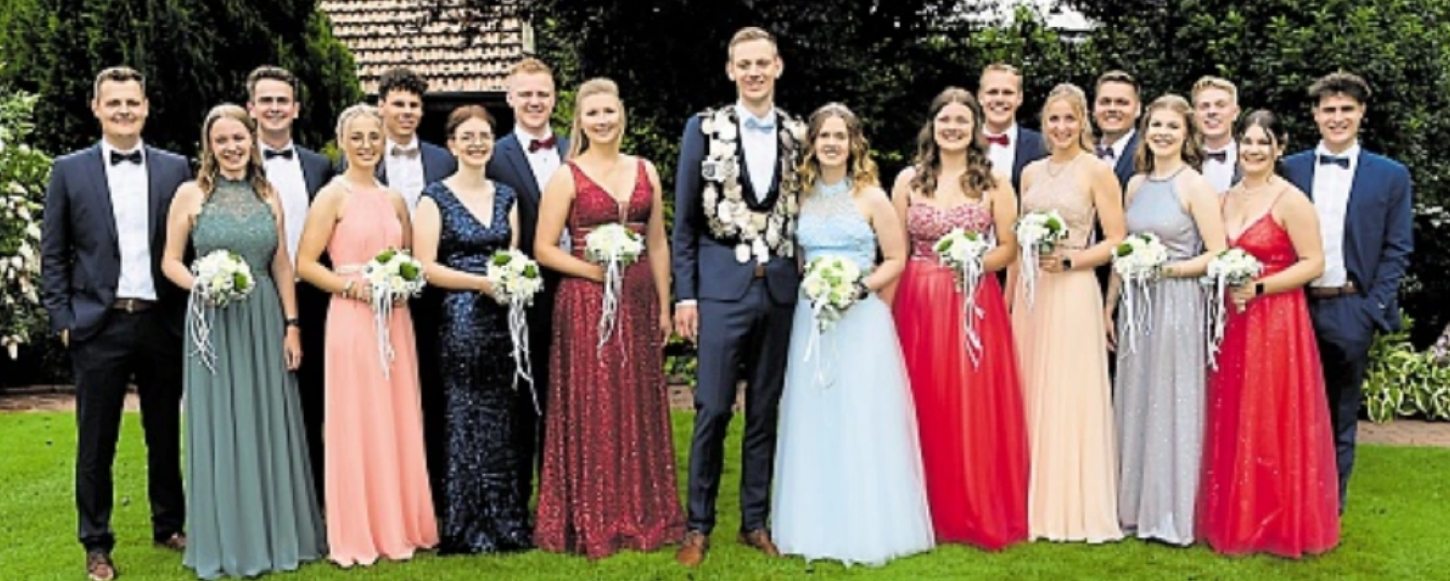 Eine Gruppe junger Erwachsene steht in einer Reihe. Sie tragen elegante Abendkleidungen und halten Blumensträuße. Die Atmosphäre ist feierlich, umgeben von Gartenlandschaft.