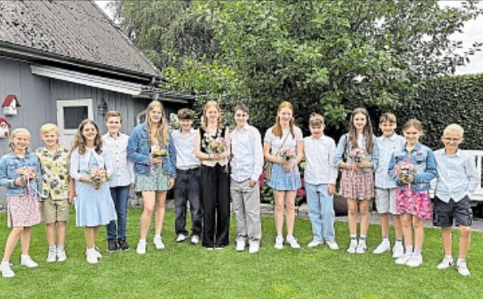 Gruppe von Kindern steht auf einer Wiese vor einem Haus; sie halten bunte Blumensträuße und tragen festliche Kleidung, einige in Jeansjacken.